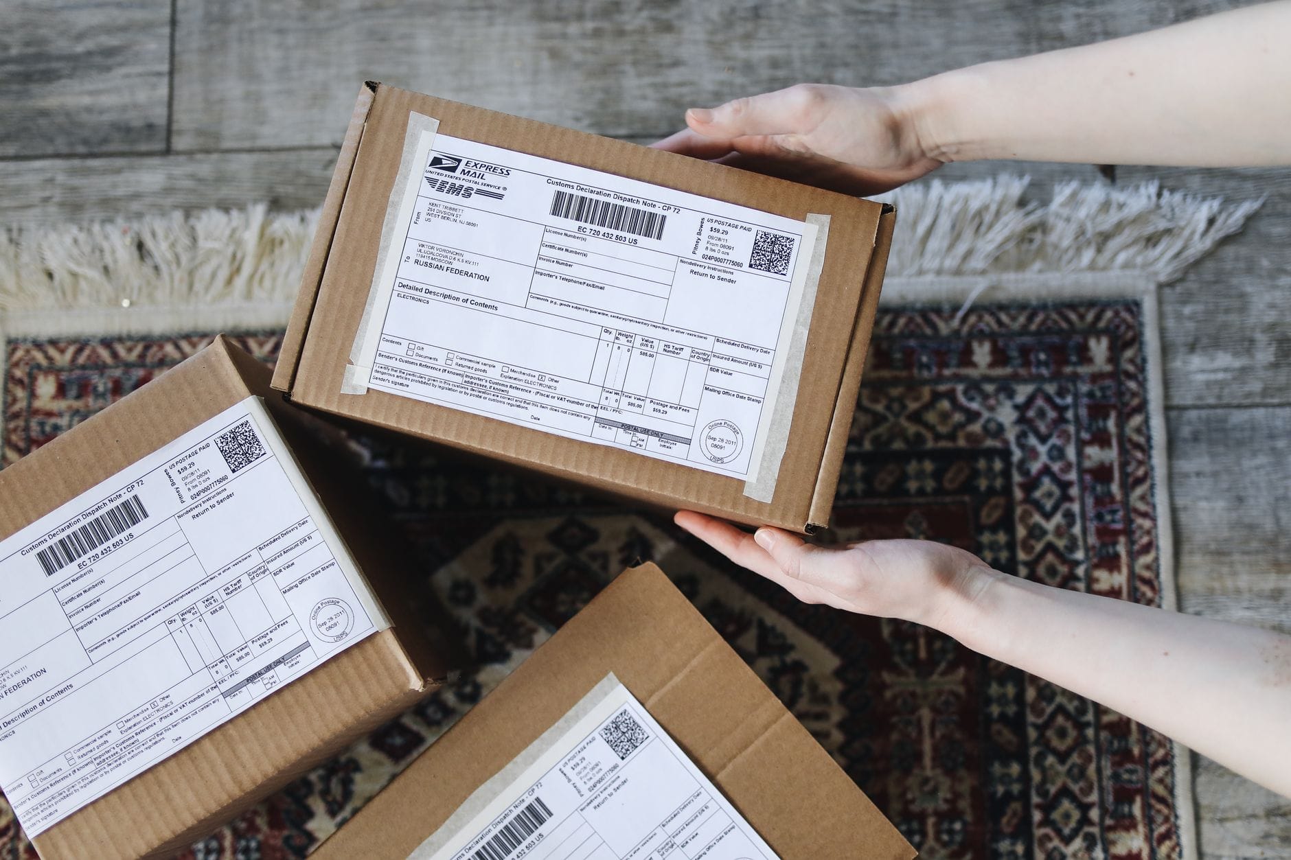 Dłonie trzymają kartonowe paczki e-commerce z etykietami na dywanie